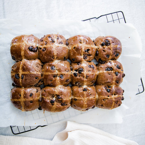 One dozen gluten free hot cross buns baked by Alternative Bread Company. Sitting on baking paper and black square rack on a white linen table cloth 